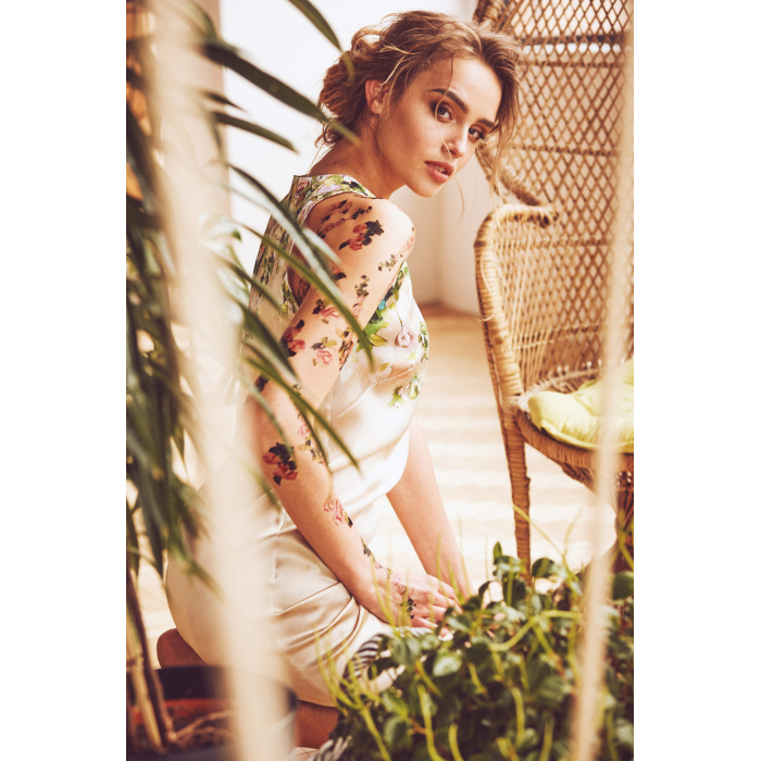 Sunny portrait of amazing young sexy woman with updo hairstyle and flower tattoos on her arms and shoulders wearing silk floral dress in studio with plants and straw chair